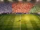 Floodlit stadium with packed colourful stands and pitch centre-spot, evening group-stage atmosphere.
