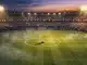 Floodlit stadium pitch at dusk with centre circle and flags in stands; pre-match atmosphere.