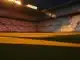 Floodlit stadium at dusk with red-white and blue-white coloured stands, dramatic shadows across empty pitch.