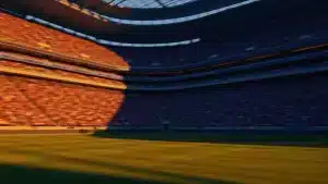 Floodlit stadium at golden hour with packed stands and dramatic shadow patterns across the pitch during a semi-final match.