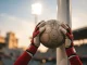 Goalkeeper gloves holding football against stadium goalpost with Arsenal colors