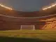 Floodlit stadium packed with crowds in red and yellow colours, dramatic shadows across empty pitch at dusk.