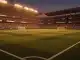 Floodlit stadium at dusk with packed crowds, red-white-blue and black-yellow-red flag colour palette, dramatic shadow patterns on pitch.