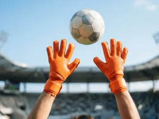 Orange goalkeeper gloves reaching up to catch a football against blue sky in stadium