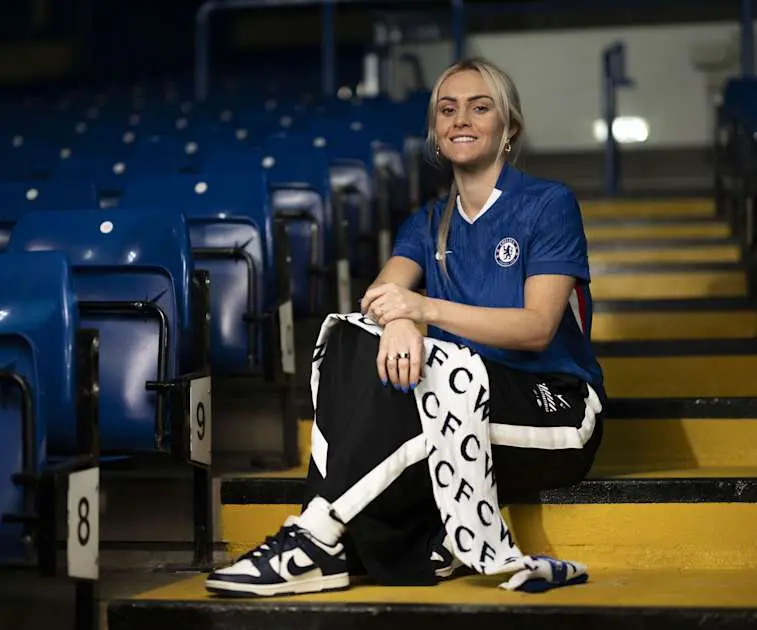 Ellie Carpenter sitting on stadium stairs wearing a blue Chelsea FC Women's jersey and team tracksuit pants.